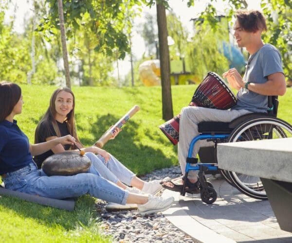Happy handicapped man on a wheelchair spending time with friends playing live instrumental music outdoors. Concept of social life, friendship, possibilities, inclusion, diversity.