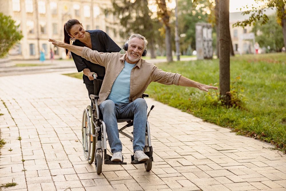 Joyful mature disabled man in wheelchair wearing headphones having fun during a walk in the city assisted by lovely young nurse